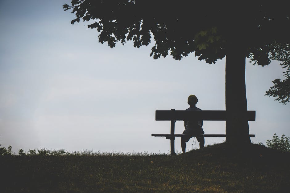 Criança sentada sozinha em um banco, embaixo de uma árvore.