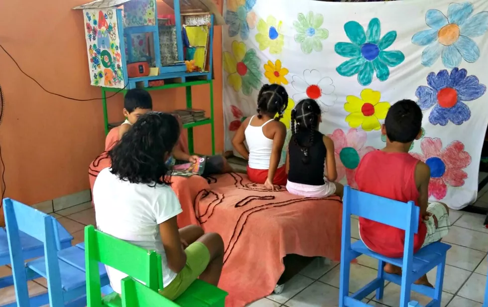 Crianças e adolescentes sentados de costa, olhando para a frente o que parece ser uma cortina improvisada com lençol para um teatro ou apresentação