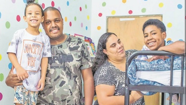 Quatro pessoas, um pai e uma mãe ao centro e duas crianças, uma em cada extremidade da imagem, sorrindo